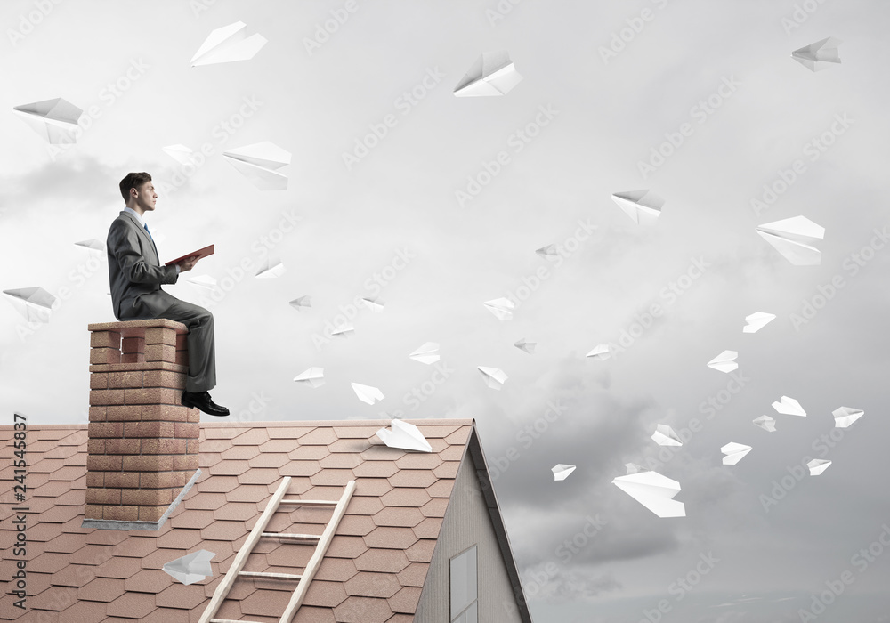 Man on brick roof reading book and paper planes flying in air Man on brick roof reading book and paper planes flying in air