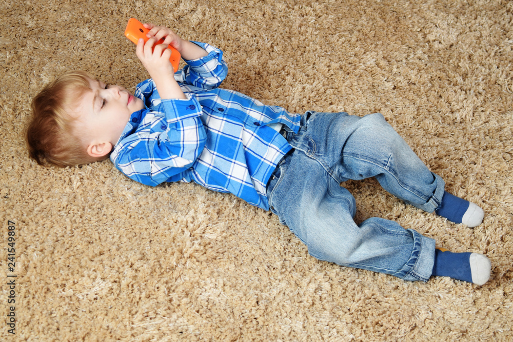 Naklejka premium A little boy is lying on the carpet and playing in the phone. Little boy using smartphone.