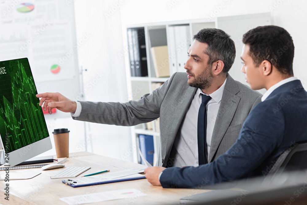 Fototapeta premium bearded businessman showing desktop computer with business graphs to young colleague at workplace