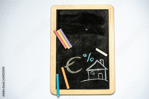 house, EURO currency sign and percentages drawn with chalk on a blackboard, next to colorful chalk on a white background, with space for text