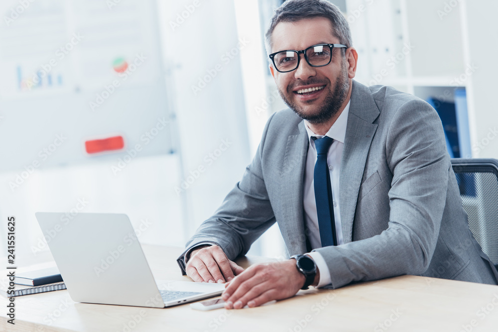 cheerful businessman in eyeglasses using laptop and smiling at camera in office
