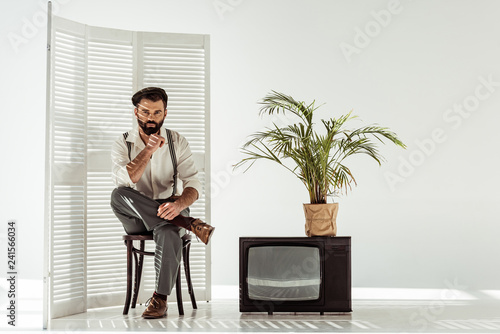 handsome bearded man sitting on chair near folding screen and tv in white room