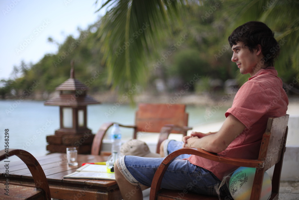 Handsome young man in sits in beach cafe by sea has vacation time. Travel concept.