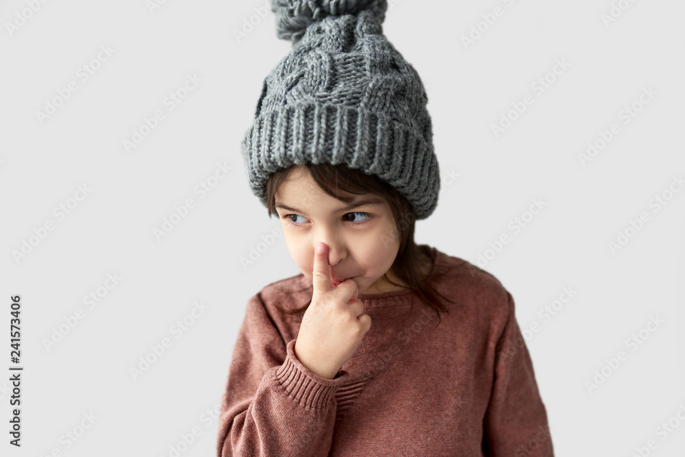 Closeup horizontal portrait of cute little girl in the winter warm gray hat, wearing sweater touching her nose, posing on a white studio background. Closeup horizontal portrait of cute little girl in the winter warm gray hat, wearing sweater touching her nose, posing on a white studio background.