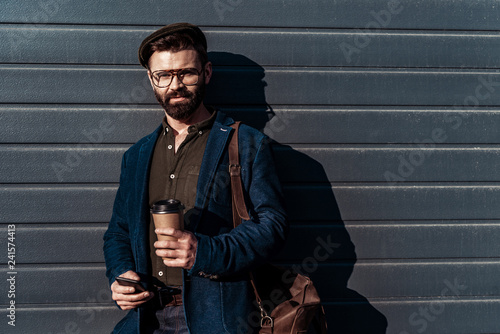 handsome bearded man in glasses and cap holding paper cup and smartphone