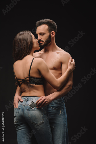 handsome shirtless man hugging girlfriend and holding condom isolated on black