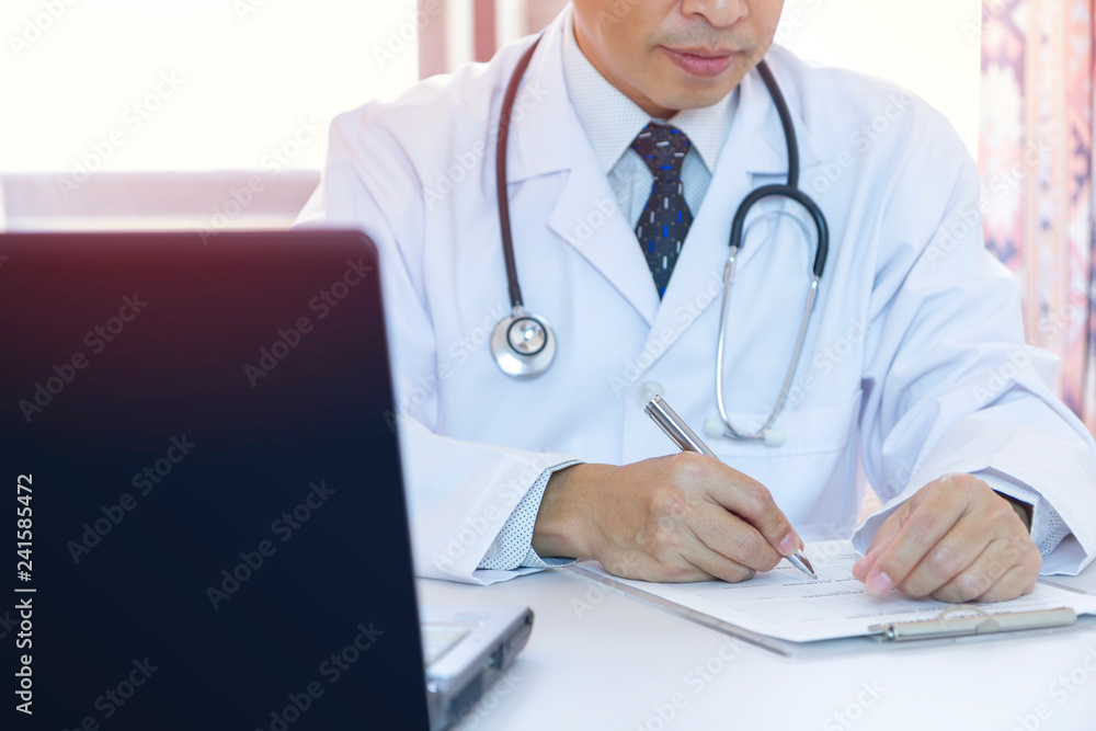 Male doctor with stethoscope writing down on a patient record in front of laptop computer notebook in a hospital office. Side copy space included. Male doctor with stethoscope writing down on a patient record in front of laptop computer notebook in a hospital office. Side copy space included.