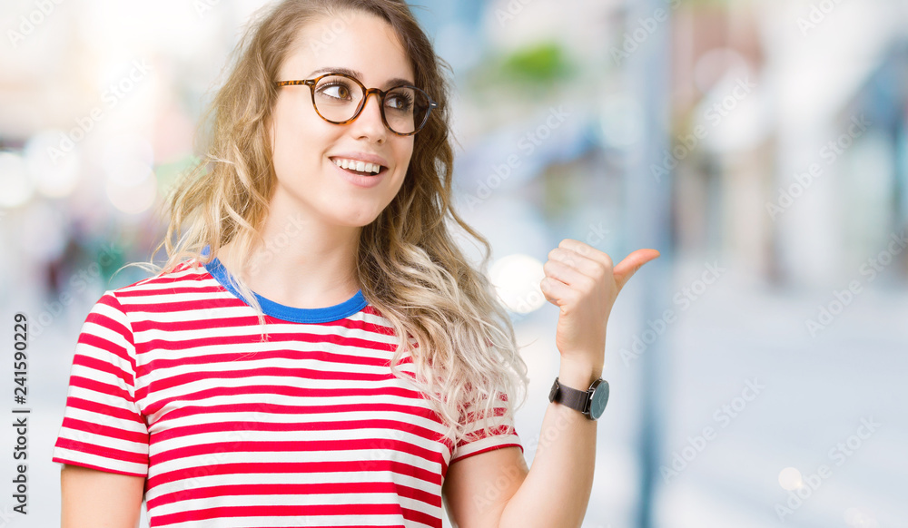Beautiful young blonde woman wearing glasses over isolated background smiling with happy face looking and pointing to the side with thumb up.