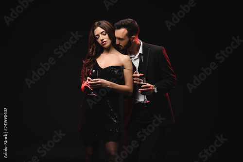 sensual couple in black dress and tuxedo holding glasses with red wine isolated on black