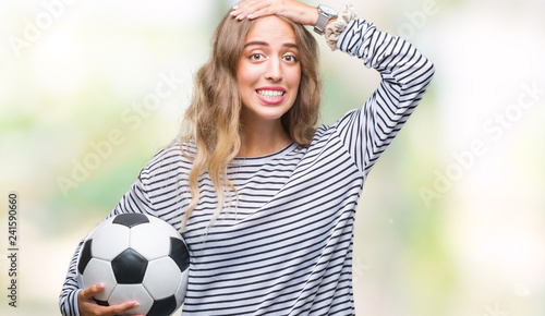 Beautiful young blonde woman holding soccer football ball over isolated background stressed with hand on head, shocked with shame and surprise face, angry and frustrated. Fear and upset for mistake. © Krakenimages.com