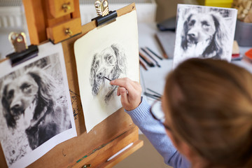 Rear View Of Female Teenage Artist Sitting At Easel Drawing Picture Of Dog From Photograph In Charcoal