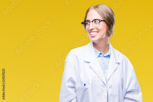 Beautiful young blonde doctor woman wearing white coat over isolated background looking away to side with smile on face, natural expression. Laughing confident.