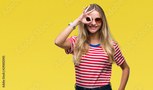 Young beautiful blonde woman wearing sunglasses over isolated background doing ok gesture with hand smiling, eye looking through fingers with happy face.