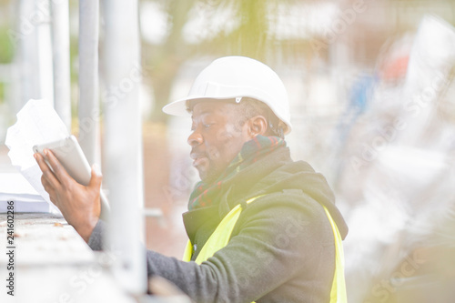 African American engineer wearing safety helmet and jacket checking documents on tablet computer on construction site