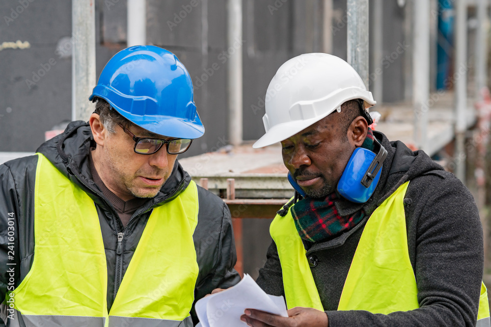 Civil engineers with hardhat and yellow jacket checking technical drawings and office blueprints among scaffolding on construction site Civil engineers with hardhat and yellow jacket checking technical drawings and office blueprints among scaffolding on construction site