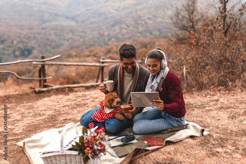 Couple at picnic sitting on the blanket and using tablet. Woman holding tablet while man holding tea and dog in his lap. In front of them basket. Autumn time. Couple at picnic sitting on the blanket and using tablet. Woman holding tablet while man holding tea and dog in his lap. In front of them basket. Autumn time.