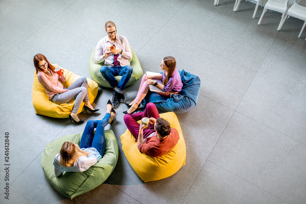 Group of a young coworkers sitting together on the colorful poufs resting during the coffee break in the office, view from above - obrazy, fototapety, plakaty 