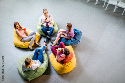 Group of a young coworkers sitting together on the colorful poufs resting during the coffee break in the office, view from above