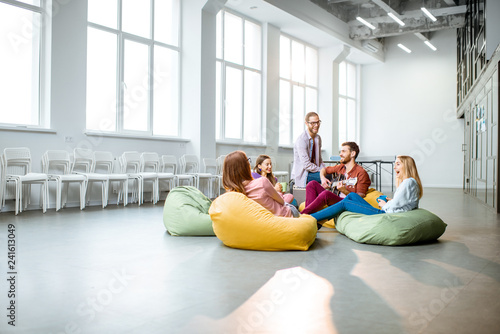 Group of a young coworkers having fun sitting on the colorful poufs playing guitar during the coffee break in the office, wide interior view