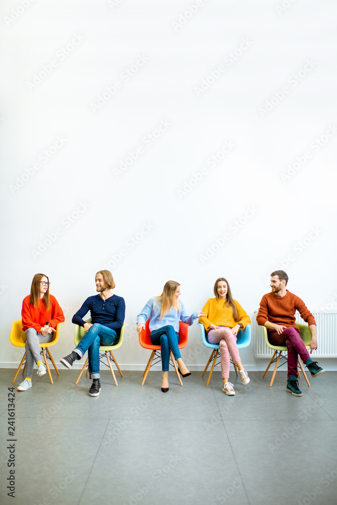 Portrait of a young people dressed casually sitting in a row on the colorful chairs on the white wall background