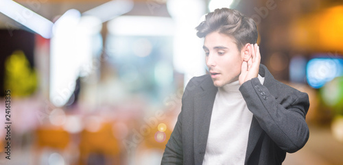 Young elegant man wearing winter coat over isolated background smiling with hand over ear listening an hearing to rumor or gossip. Deafness concept.