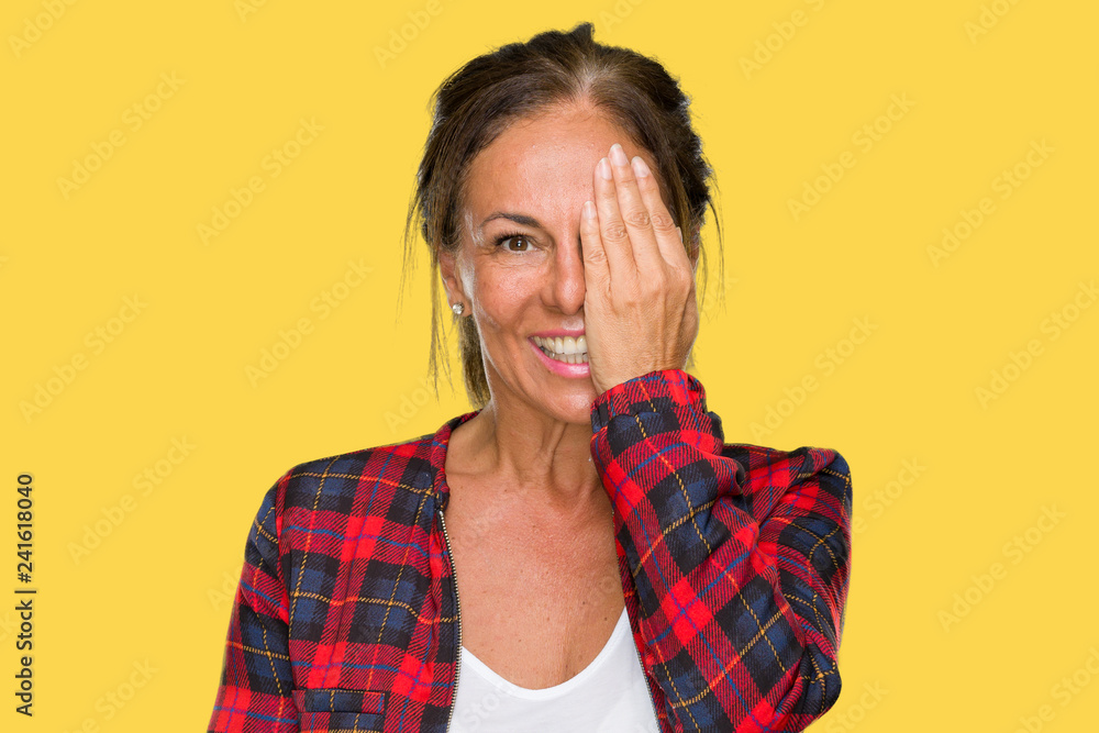 Middle age adult woman wearing casual jacket over isolated background covering one eye with hand with confident smile on face and surprise emotion. Middle age adult woman wearing casual jacket over isolated background covering one eye with hand with confident smile on face and surprise emotion.