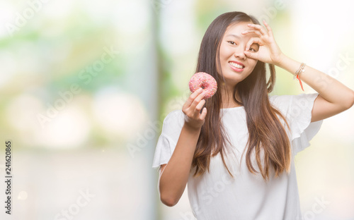 Young asian woman eating donut over isolated background with happy face smiling doing ok sign with hand on eye looking through fingers