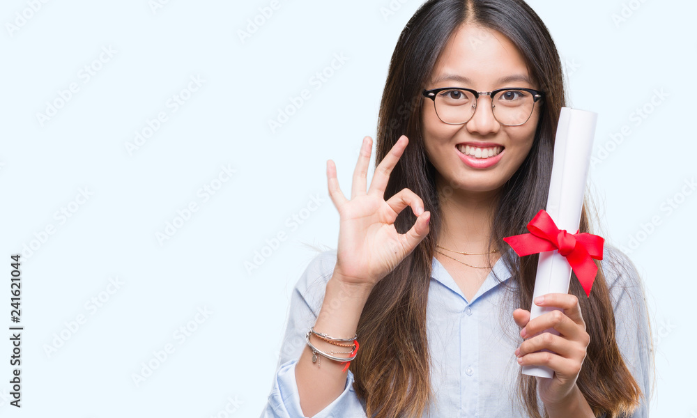 Naklejka premium Young asian woman holding degree over isolated background doing ok sign with fingers, excellent symbol