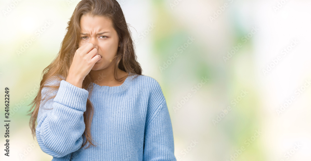 Young beautiful brunette woman wearing blue winter sweater over isolated background smelling something stinky and disgusting, intolerable smell, holding breath with fingers on nose