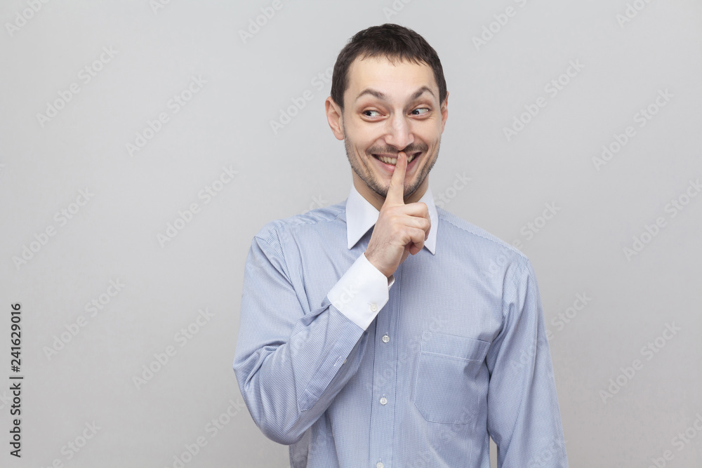 Shh, this is secret. Funny handsome bristle businessman in classic light blue shirt standing with silence alarm and looking away with toothy smile. indoor studio shot, isolated on grey background. Shh, this is secret. Funny handsome bristle businessman in classic light blue shirt standing with silence alarm and looking away with toothy smile. indoor studio shot, isolated on grey background.