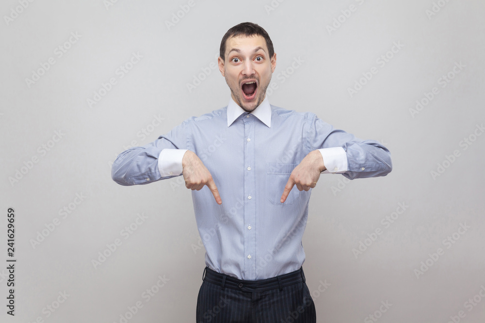 Portrait of surprised handsome bristle businessman in classic blue shirt standing and pointing at down copyspace and looking at camera with amazed face. indoor studio shot, isolated on grey background Portrait of surprised handsome bristle businessman in classic blue shirt standing and pointing at down copyspace and looking at camera with amazed face. indoor studio shot, isolated on grey background