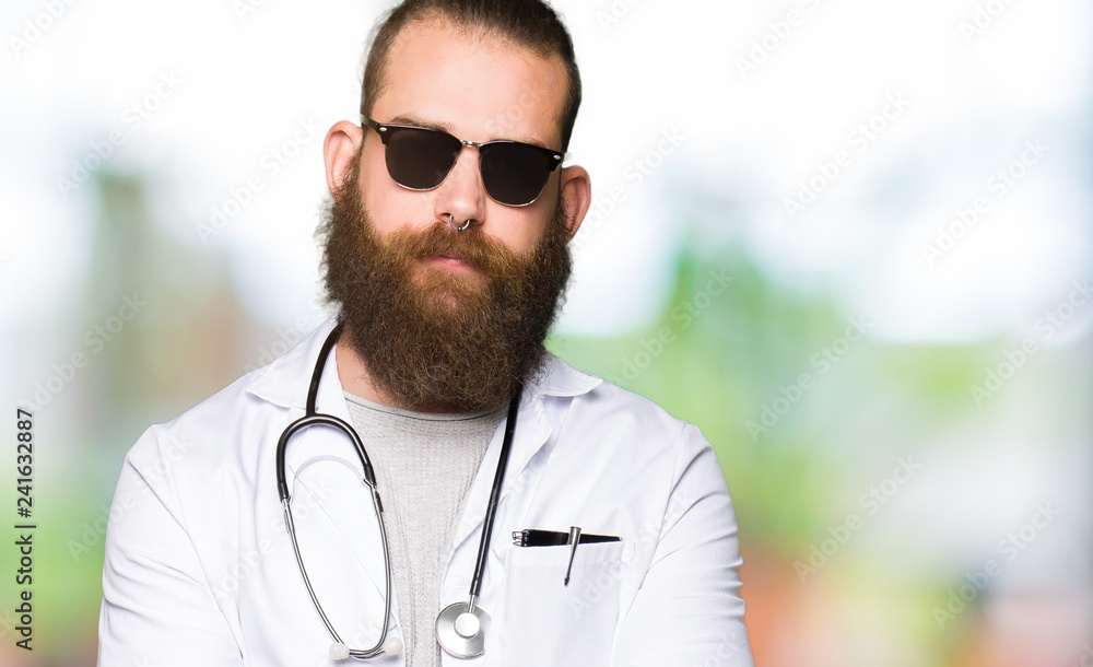 Young blond doctor man with beard wearing sunglasses skeptic and nervous, disapproving expression on face with crossed arms. Negative person. Young blond doctor man with beard wearing sunglasses skeptic and nervous, disapproving expression on face with crossed arms. Negative person.