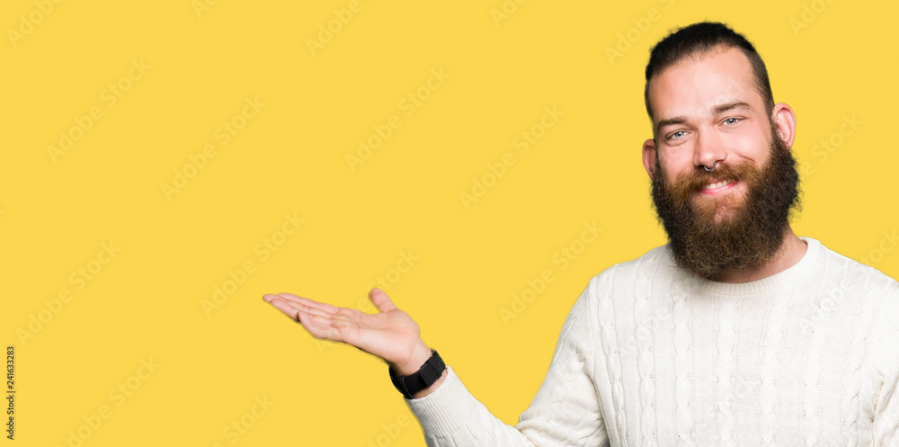 Young hipster man wearing winter sweater smiling cheerful presenting and pointing with palm of hand looking at the camera. Young hipster man wearing winter sweater smiling cheerful presenting and pointing with palm of hand looking at the camera.