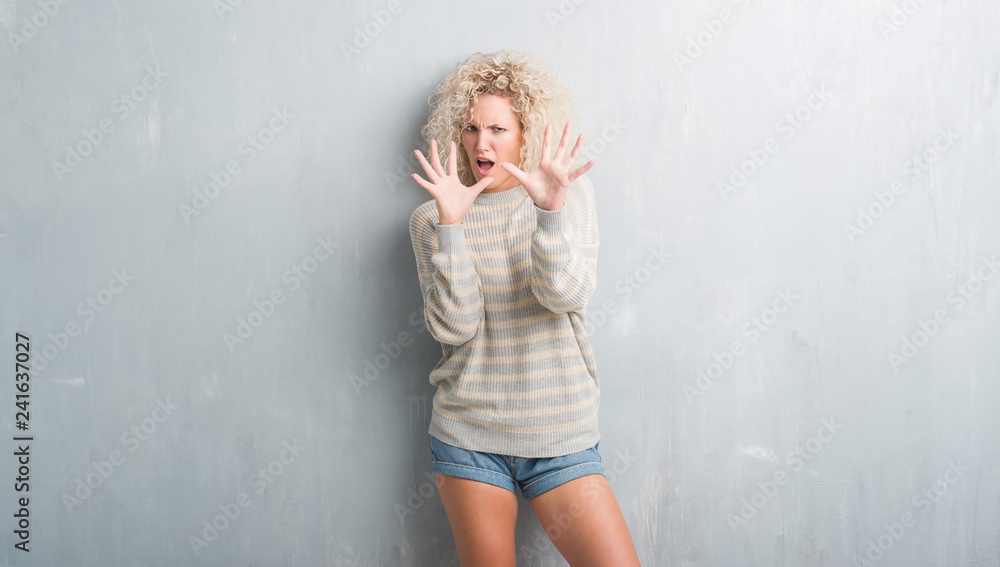 Young blonde woman with curly hair over grunge grey background afraid and terrified with fear expression stop gesture with hands, shouting in shock. Panic concept.