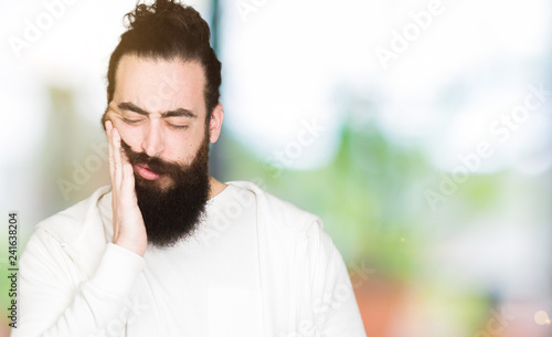 Young man with long hair and beard wearing sporty sweatshirt touching mouth with hand with painful expression because of toothache or dental illness on teeth. Dentist concept.