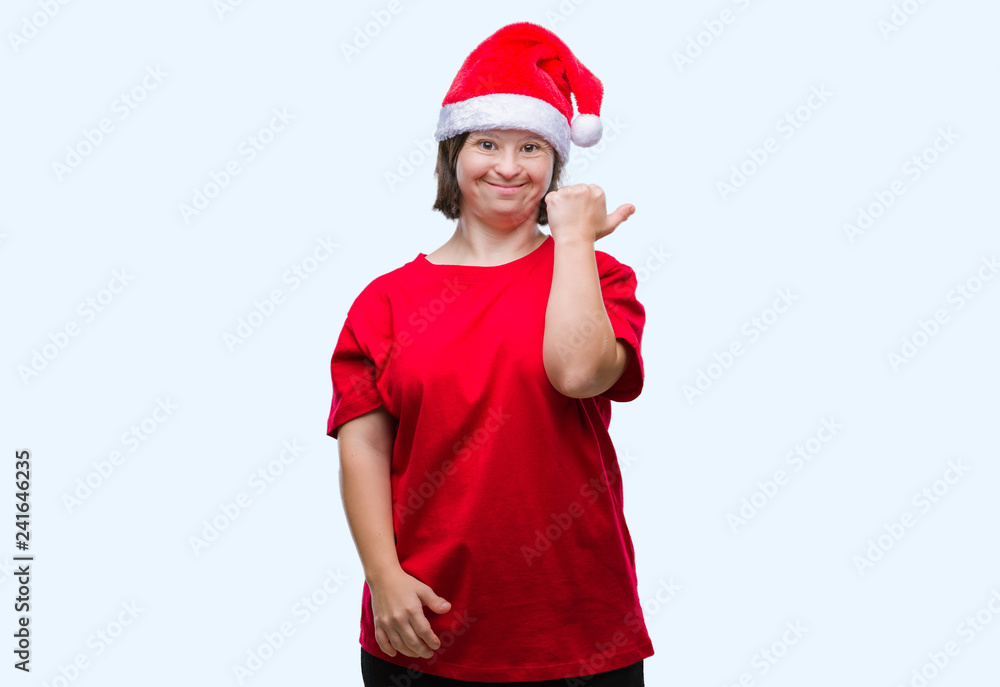 Young adult woman with down syndrome wearing christmas hat over isolated background smiling with happy face looking and pointing to the side with thumb up. Young adult woman with down syndrome wearing christmas hat over isolated background smiling with happy face looking and pointing to the side with thumb up.