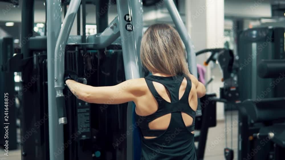 Young fit woman doing shoulders exercise using training machine in gym. View from back. Slowmotion