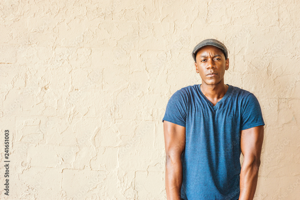 Portrait of Young African American Man in New York. A black guy wearing blue V neck T shirt, flat cap, straightening arms, shrugging shoulders, standing by painted wall on street, looking, thinking.