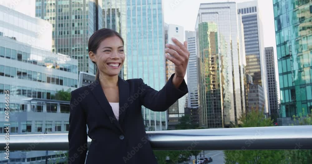 Smiling business professional waving while holding mobile phone in financial district. Young businesswoman having video call on mobile phone against office buildings. She is wearing suit. Smiling business professional waving while holding mobile phone in financial district. Young businesswoman having video call on mobile phone against office buildings. She is wearing suit.