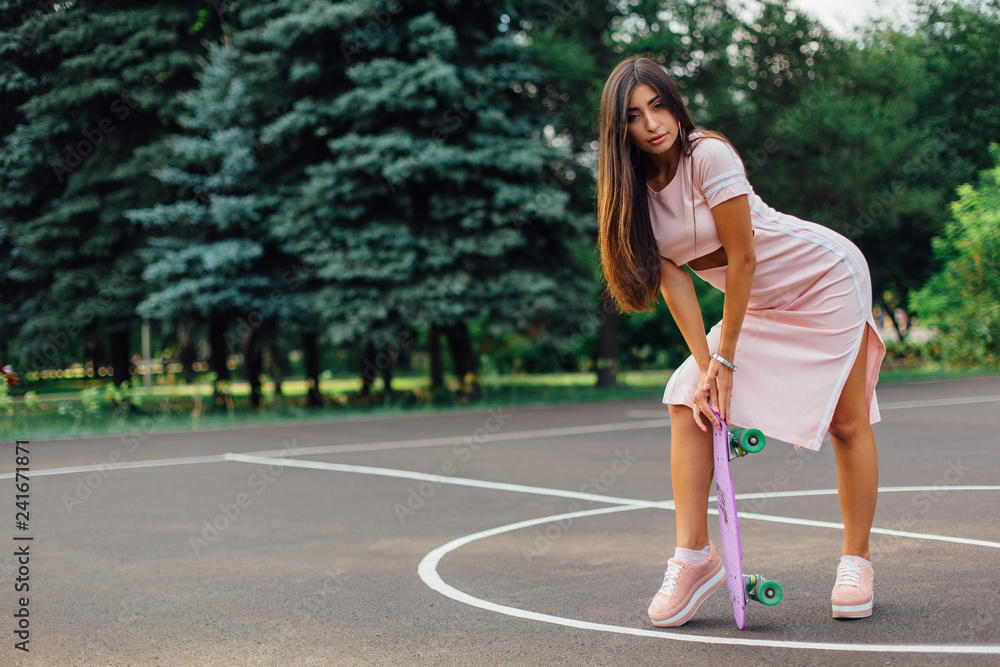 Portrait of a smiling charming brunette female holding her skateboard on a basketball court. Portrait of a smiling charming brunette female holding her skateboard on a basketball court.