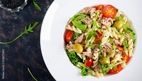 Pasta salad with tuna, tomatoes, olives, cucumber, sweet pepper and arugula on rustic background . Top view