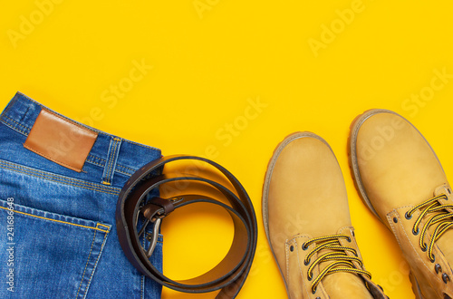 Fashionable concept. Men's casual wear, yellow work boots from natural nubuck leather, blue jeans and brown belt on bright yellow background top view flat lay copy space. Trendy casual shoes clothes