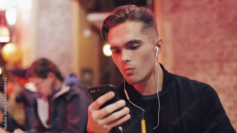 Handsome young man uses headphones and his smartphone for video call. He sits at the bar near the neon signs and drink coctail. Cheerful mood, modern communication, technologies, devices Handsome young man uses headphones and his smartphone for video call. He sits at the bar near the neon signs and drink coctail. Cheerful mood, modern communication, technologies, devices