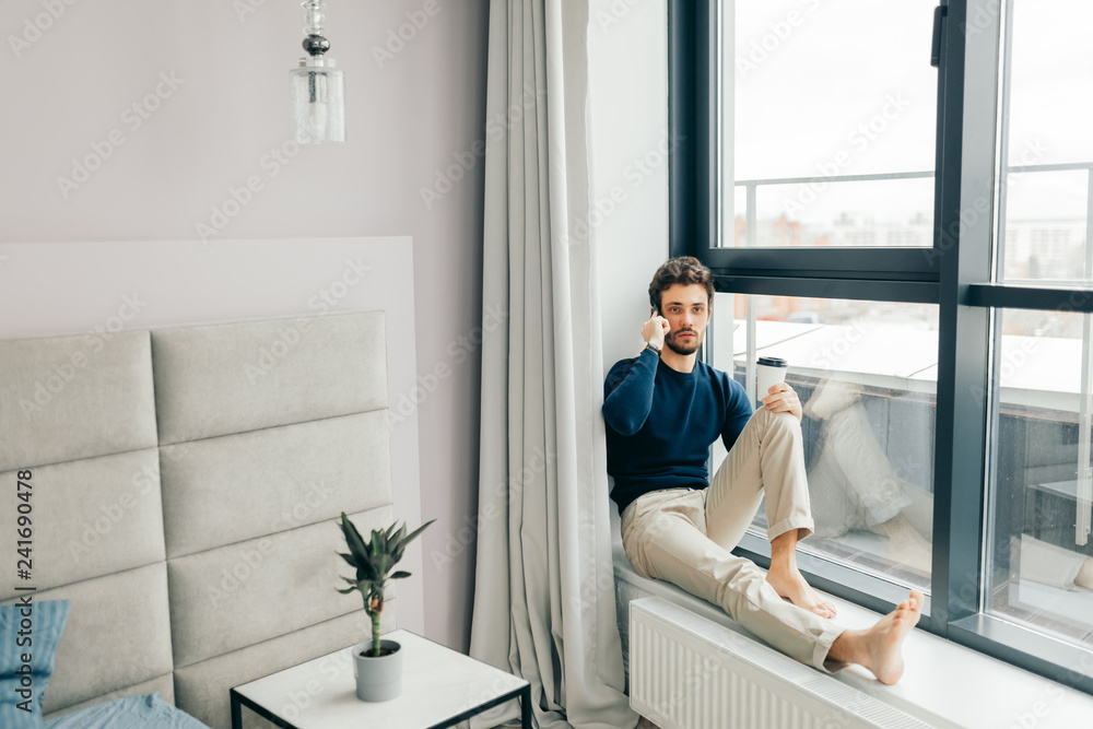 Handsome caucasian man in casual clothes speaking on phone, sits on panoramic window sillin hotel room with modern stylish interior Handsome caucasian man in casual clothes speaking on phone, sits on panoramic window sillin hotel room with modern stylish interior