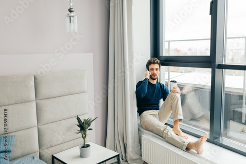 Handsome caucasian man in casual clothes speaking on phone, sits on panoramic window sillin hotel room with modern stylish interior