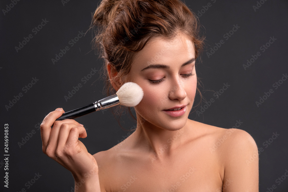 Obraz premium Beautiful brunette caucasian young woman prepare herself, applying powder on her cheeks with a brush. Clean, fresh, natural, flawless skin. Soft smile on her face. Close up on a neutral background