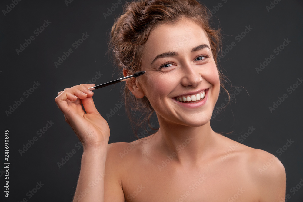 Fototapeta premium Beautiful brunette caucasian young woman prepare herself, applying mascara on her eyes with a brush. Clean, fresh, natural, flawless skin. Soft smile on her face. Close up on a neutral background