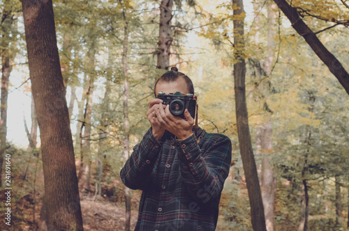 Hipster millennial young man or teenager with analog vintage photo camera makes photograph into lens in middle of old wood with sunset light, explores tourism. Hipster Travel Lifestyle concept