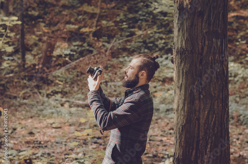 Hipster Travel Lifestyle concept: stylish bearded man, dressed in a shirt and with a hairdo Top Knot with retro photo camera in hands takes pictures in the woods