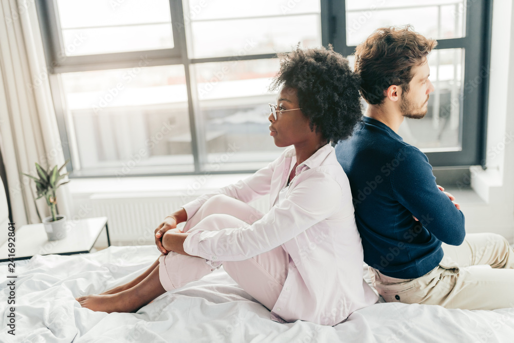 mixed race young couple sitting on bed, backside to each other, having argument with each other in bedroom, ignoring each other. mixed race young couple sitting on bed, backside to each other, having argument with each other in bedroom, ignoring each other.